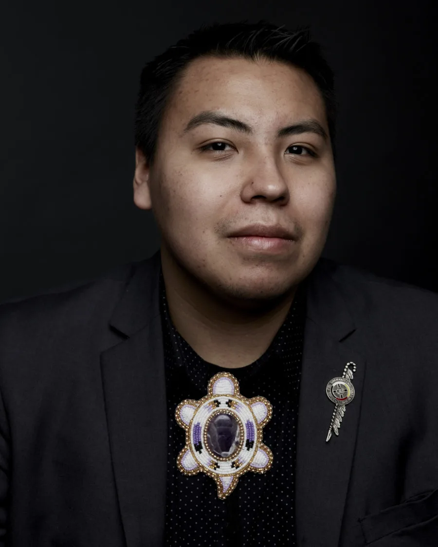 A portrait photo of a man with a light-medium skin tone wearing a black blazer and a black shirt with a white, purple, and gold beaded turtle image on the chest underneath a neck with an image in the middle in front of a solid black background. 