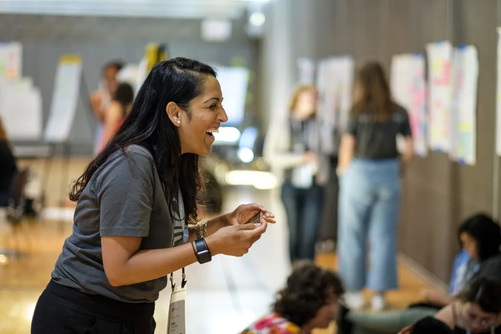 A woman with a light-medium skin tone wearing a gray shirt with a white Obama Foundation logo and black pants smiles toward the right-hand side of the photo holding a phone. In the background, there are people and white posters with pink, orange, yellow, and blue words and sticky notes blurred out.