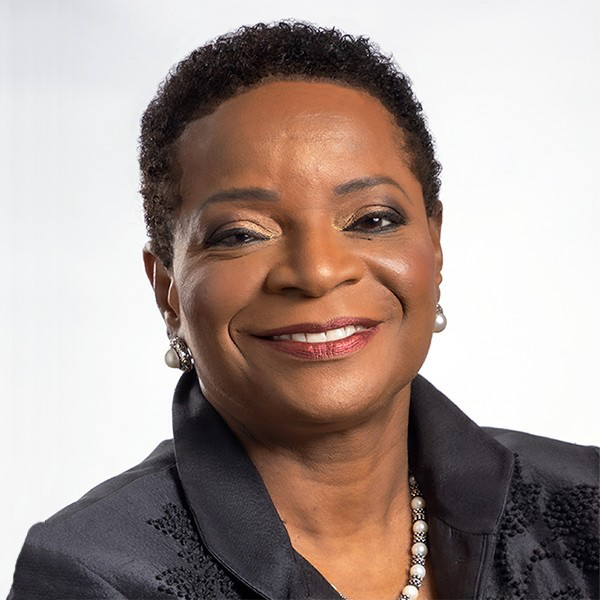 A woman with medium-dark skin tone wears a black collared jacket, pearl earrings and necklace as she smiles for the camera.