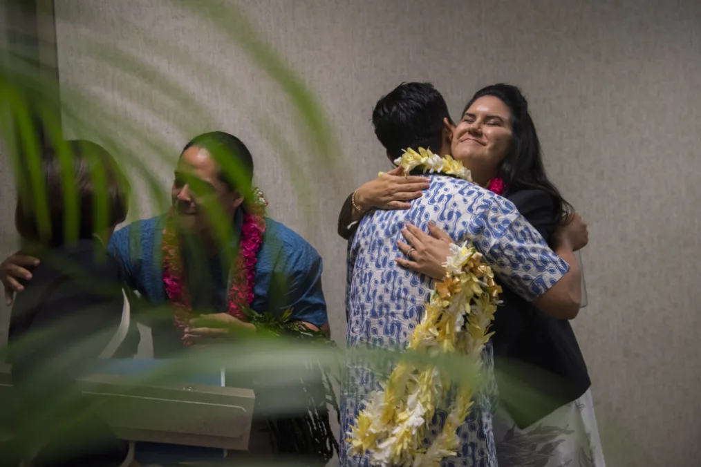 Two people embrace. The woman is holding yellow leis and the man is wearing a leis. Another pair embraces to the left.