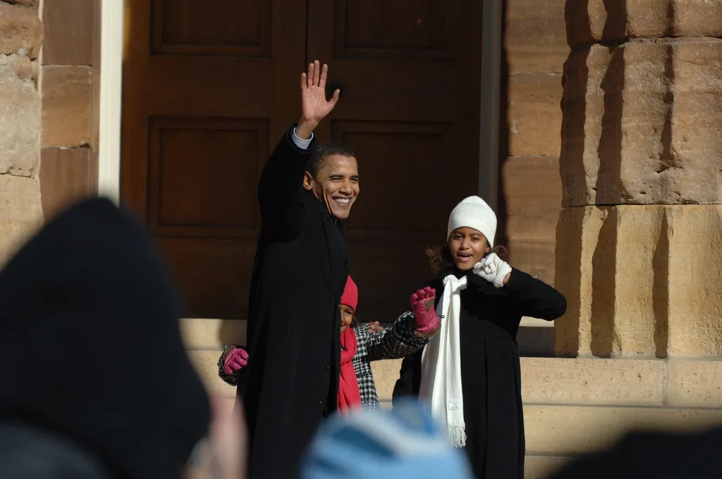 Barack, Sasha and Malia Obama wave to out-of-focus onlookers. They wear hats and scarves.