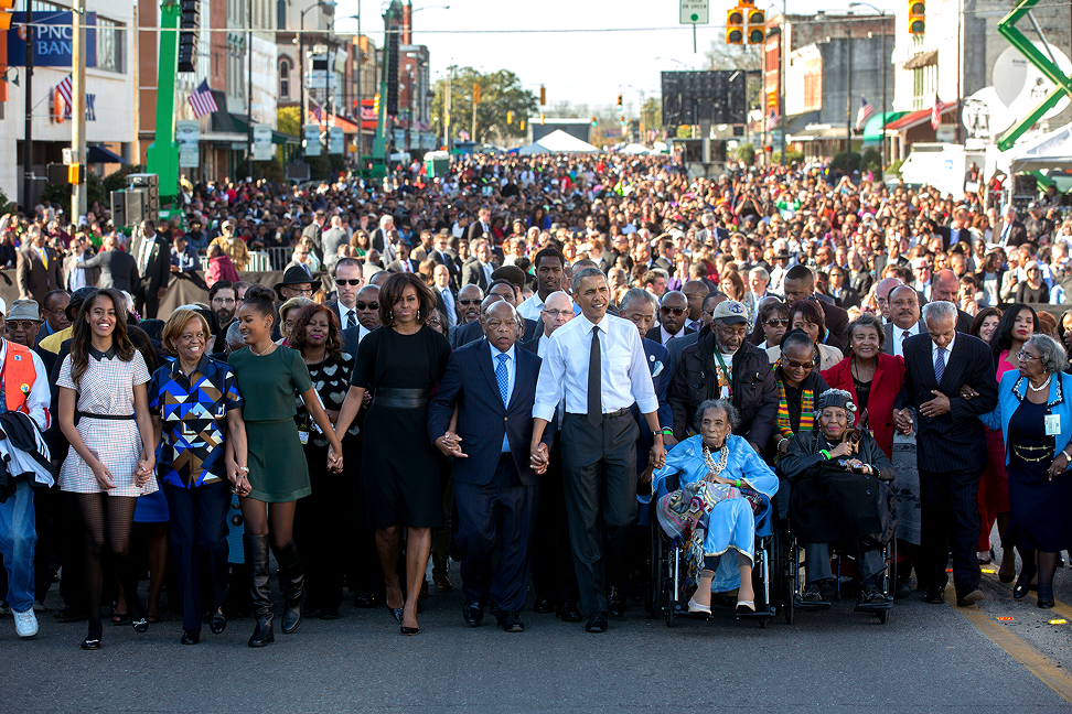 President Barack Obama, First Lady Michelle Obama and daughters Sasha and Malia wait with former President George W. Bush and former First Lady Laura Bush prior to the walking across the Edmund Pettus Bridge to commemorate the 50th Anniversary of the Selma to Montgomery civil rights marches in Selma, Alabama. There is a crowd of thousands of people behind them. 