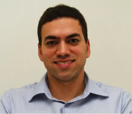 A headshot of a man with dark brown hair smiling at the camera. He wears a light blue button-up shirt.