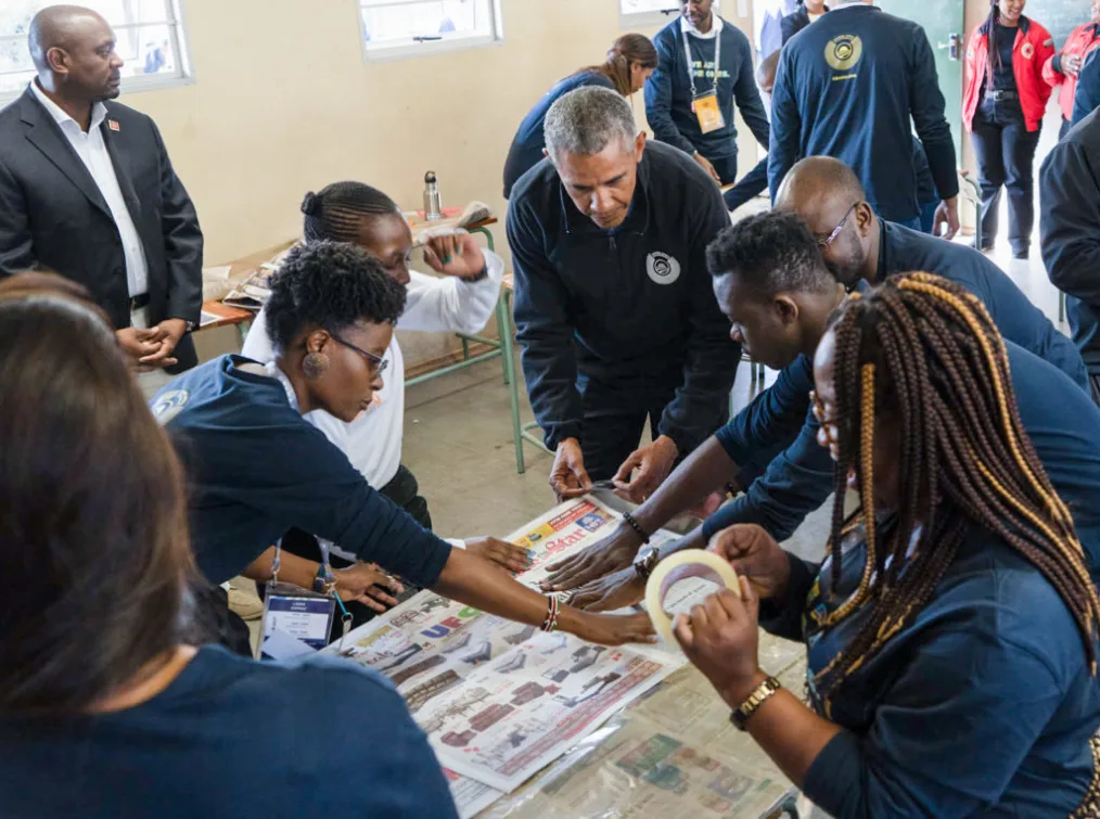 President Obama participates in an activity that involves newspaper. He works alongside a group people with deep skin tones.