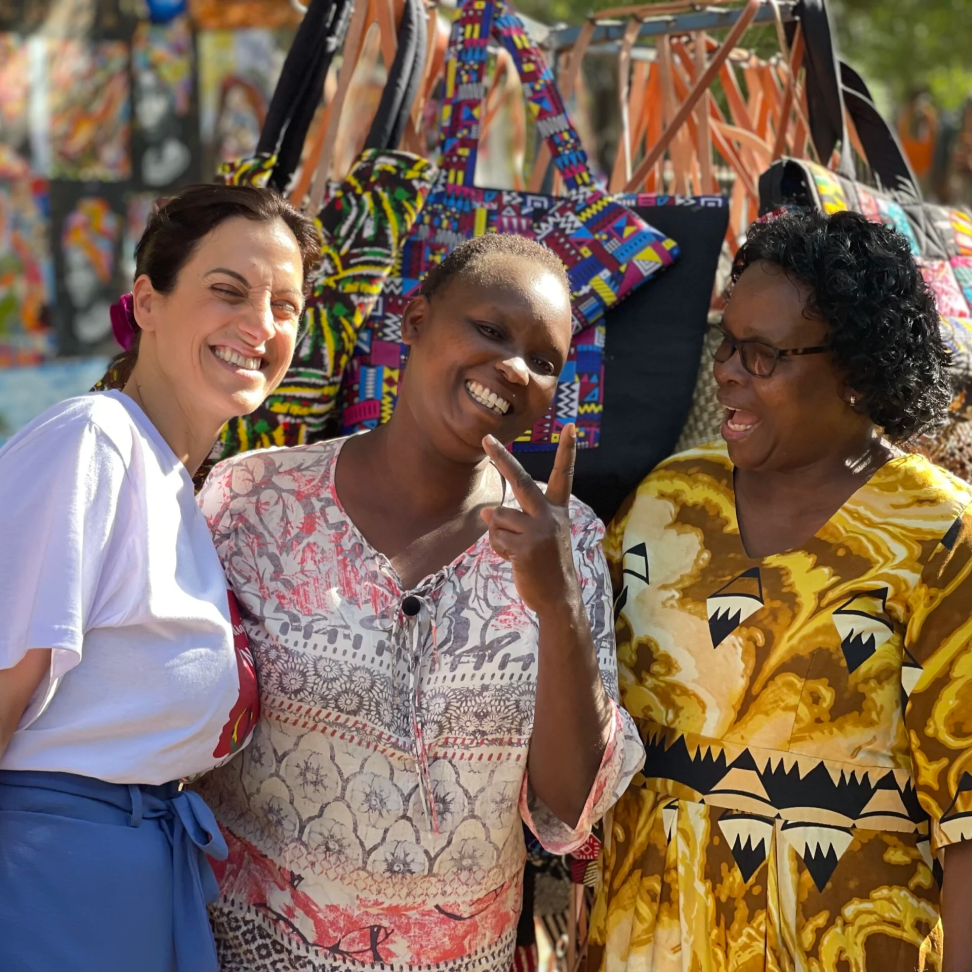 Elpida Kokkota smiles as she stands next to Jame Kalekye Mutambya, a Black woman with a deep skin tone and short hair, and Mercy Musomi, a Black woman with a deep skin tone and short curly hair. Jame holds a peace sign as they stand in front of a booth at a Naroibi cultural market. In the background is art and handmade bags. 