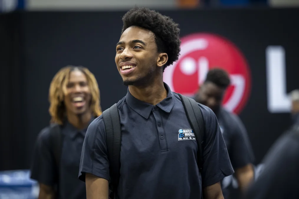 A young Black man with a medium skin tone smiles away from the camera. He has a short afro and is wearing a polo shirt that reads, “March Madness.” Two men are blurred in the background.