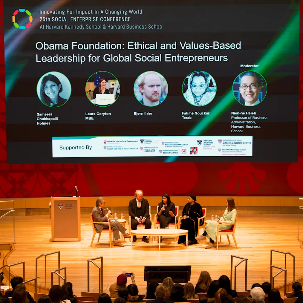 Sameera, a woman with a medium skin tone and black hair, sits on a panel at Harvard University with four others. She sits in the middle and holds a microphone. A sign in the background reads, “Obama Foundation: Ethical and Values-Based Leadership for Global Entreprenuers.” An audience is in front of them.