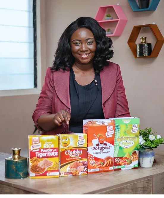 Obama Leader Vera Osei-Bonsu poses with her healthy cereal products at her office. She has a dark skin tone and shoulder length black hair. She is wearing a burgundy leather blazer and a black top.