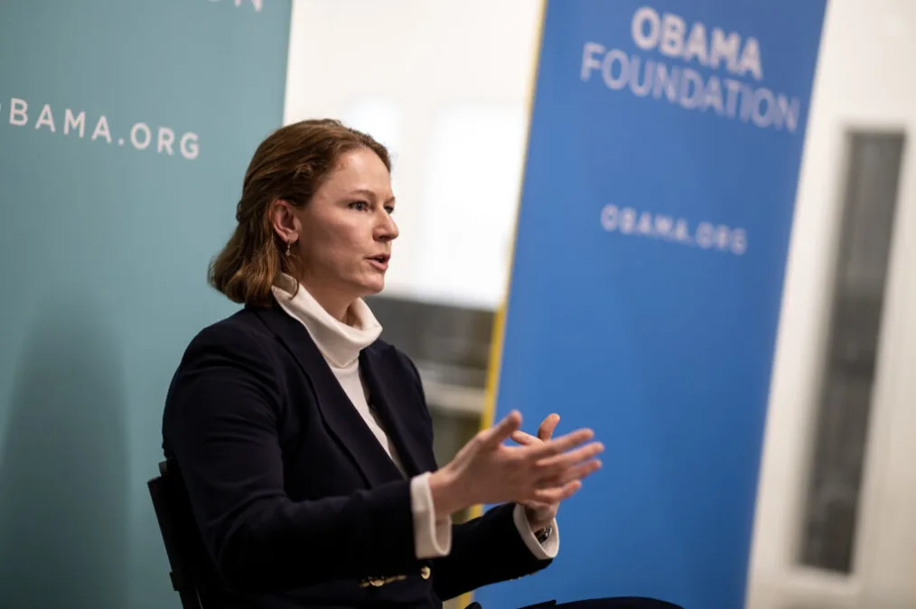 A lady with light skin, blonde-red hair in a navy blazer with white dress underneath. She is stting and explaing something to a group out of frame. In the background are Obama Foundation signs out of focus.