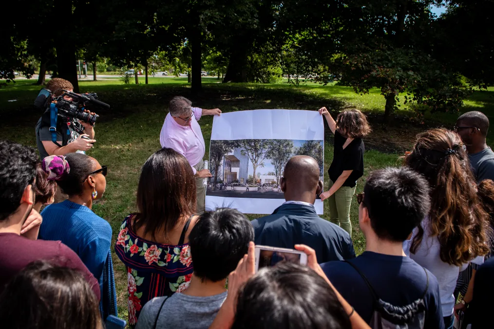 A OPC tour group looking at a image of the propected OPC site they are currently standing at. Two group leaders are holding up the image and to the left is video coverage. 