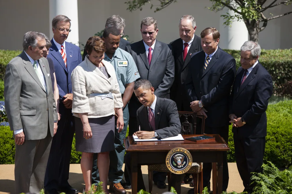 President Obama sits outdoors at a desk while signing a paper. Around him stands a group of people in various skin tones and in various business attire / uniforms 