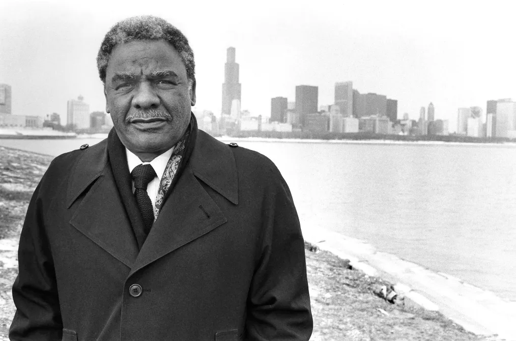 This picture shows a man with a deep skin tone in a suit and tie outside of a large city near the shore,
posing toward the camera.
