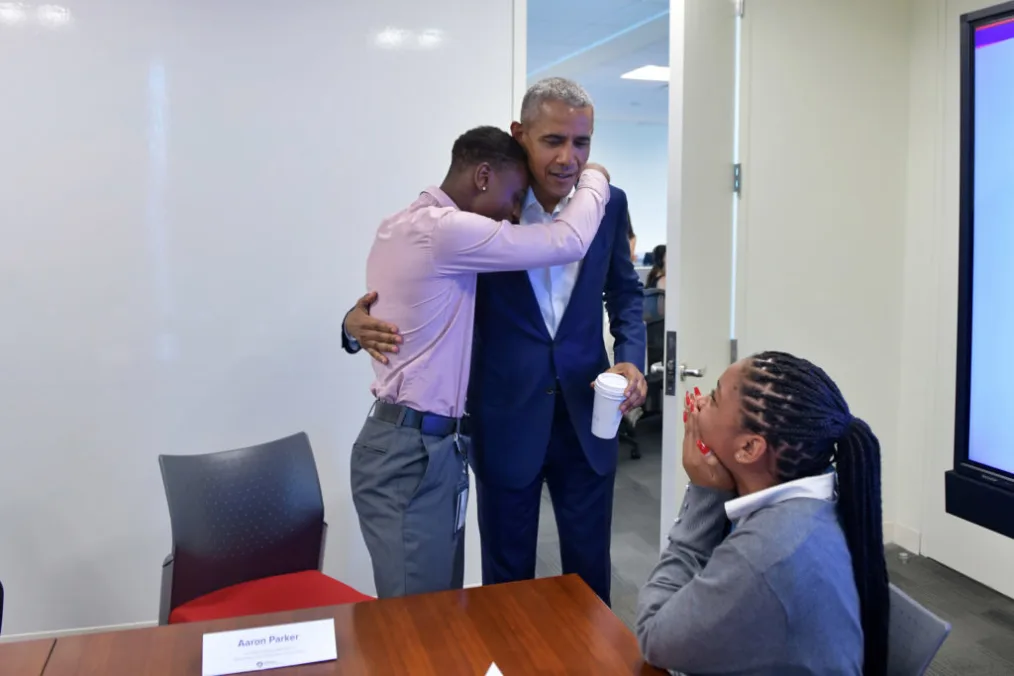 A young man hugs President Obama from the side as he holds a cup of coffee.