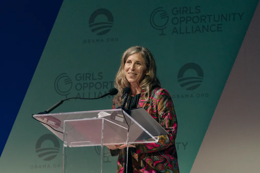 A woman with light skin and darker blonde hair stands behind a clear podium and speaks into a microphone. A sign in the background reads, “Girls Opportunity Alliance” and “Obama.org” alongside the Obama Foundation rising sun logo.