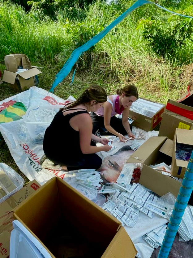 Liza Zaruba, a woman with a light skin tone and long blonde hair, sits on a tarp with another woman with a light skin tone and blonde hair, sorting medical supplies in Agbelouve, Togo.