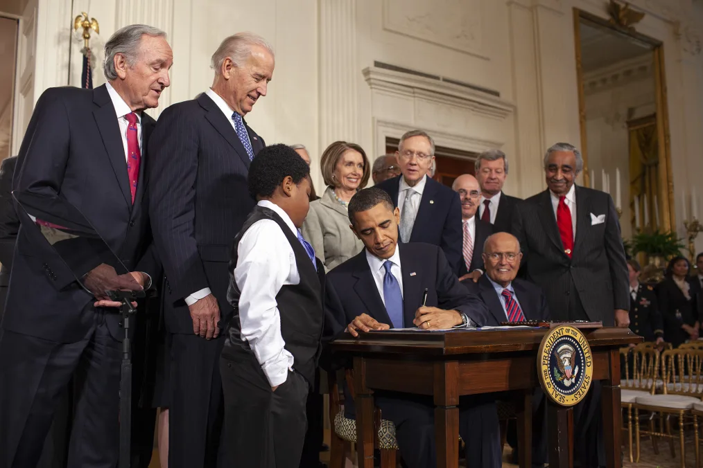 President Obama sits at a formal desk in the center of the image, glancing down as he signs a document. A young boy in a vest and tie looks on, as do Vice President Biden, Nancy Pelosi.