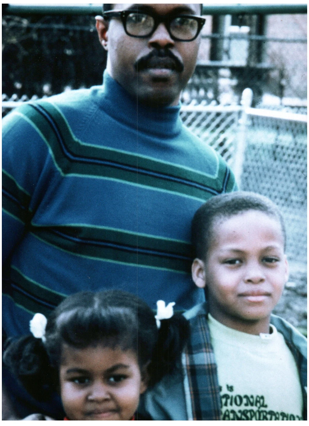 Fraser C. Robinson III, a Black man with a deep skin tone, wears a blue and green striped sweater and glasses and stands in front of a young Michelle and Craig Robinson