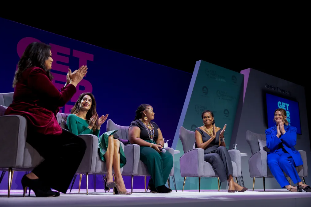 Sara Sidner, Amal Clooney, Michelle Obama, and Melinda French Gates clap for Wanjiru Wahome as they sit on a stage at the Girls Opportunity Alliance Get Her There event. The women have a range of skin tones and wear combinations of pantsuits, skirts, blazers, and other dresswear. Multiple signs in the background read, “Get Her There.”