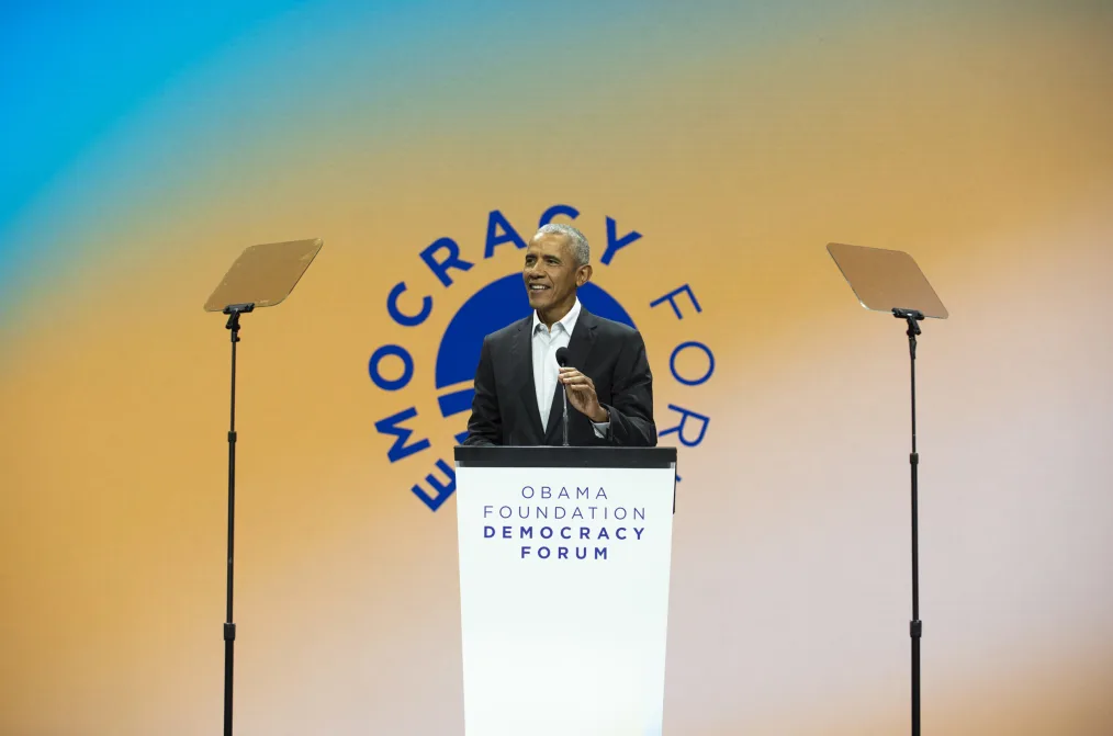President Obama stands behind a podium as he speaks at the Obama Foundation Democracy Forum. The president wears a black suit and white collared shirt. In the background is a blue, green, and yellow gradient graphic that reads, “Democracy Forum.”
