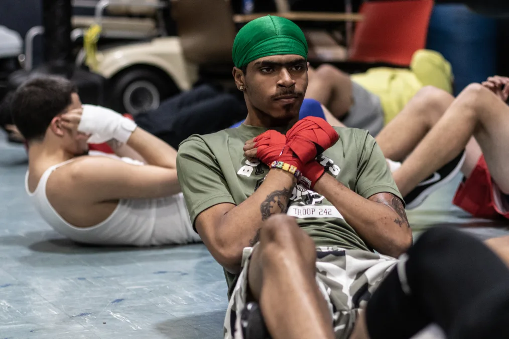 A group of young men are doing sit ups in a gym. A young man with medium skin tone is in focus in the photo. He has a light green durag on, a scrunched face, an olive shirt, and camo shorts. Wrapped around his hands is red boxing tape.