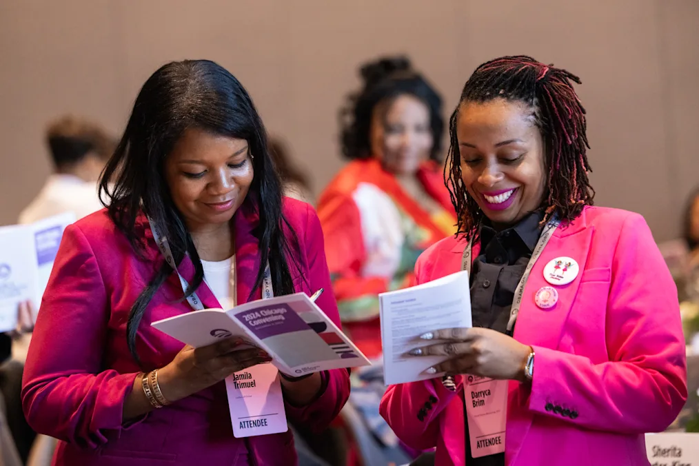 Two girl-serving leaders smile as they share notes during the Alliance’s Chicago convening. Both have medium dark skin tones and are wearing pink blazers with attendee credentials.