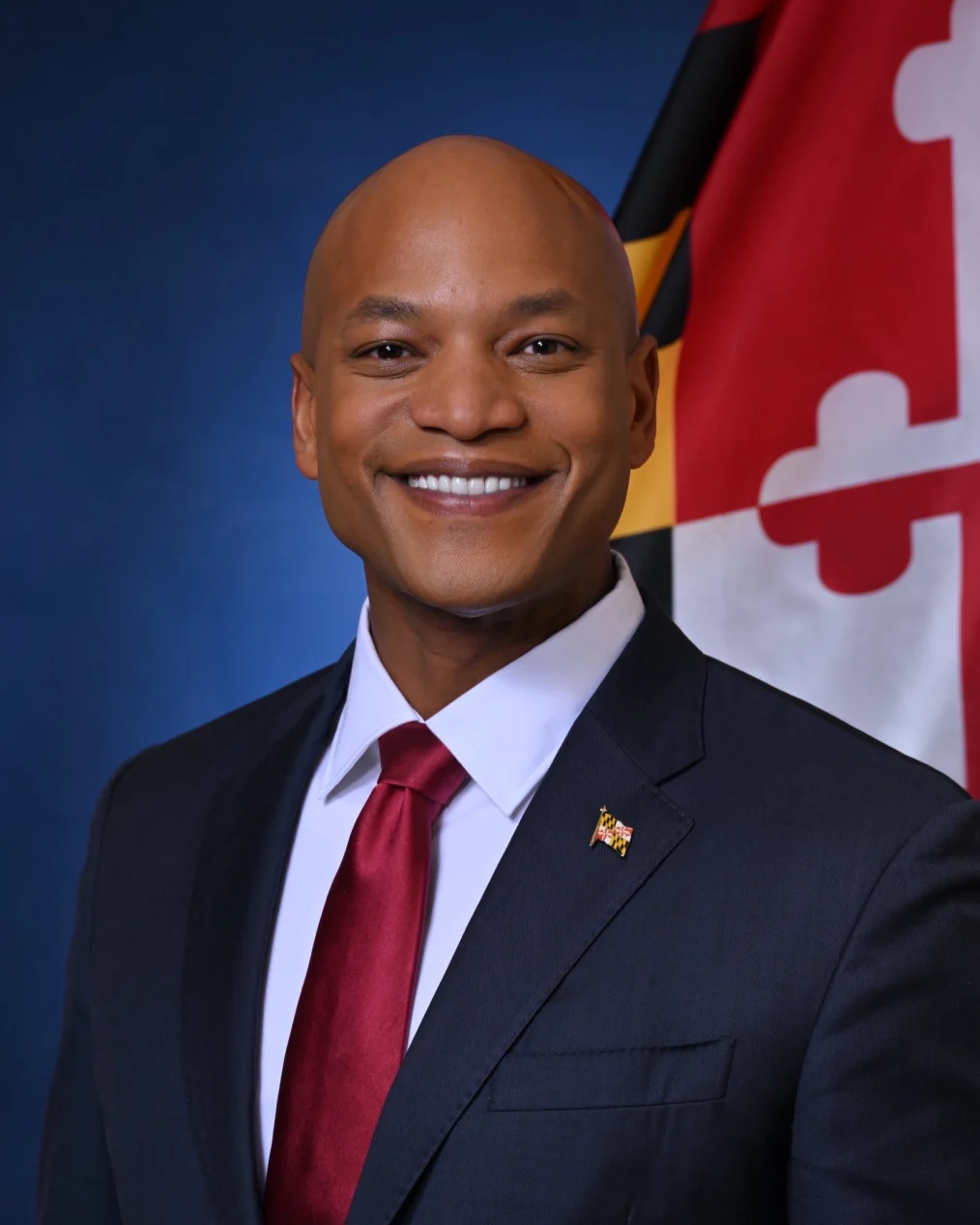 A Black man with a bald head smiles, showing his top row of teeth. He wears a black suit jacket over a white collared shirt with a red tie and a flag lapel pin. Behind him is part of the Maryland state flag, with red, yellow, white and black geometric shapes.