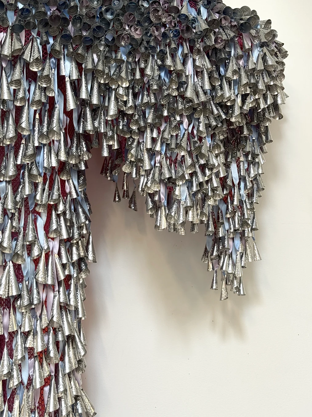 An image is a close-up of of Marie Watt's installation Sky Dances Light. The installation is made up of tin jingles attached to a white wall. The jingles are silver cones. There are thousands of them. In between the jingles are straps of pink and blue fabric.