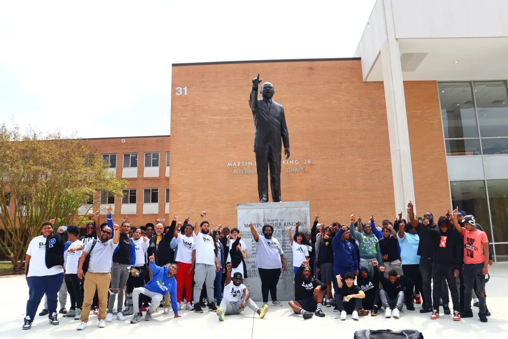 DeKevious Wilson, a Black man with a deep skin tone, points up as he stands in the middle in front of a Martin Luther King Jr. Statue. He is surrounded by a group of young men that range from light to deep skin tones. All are pointing toward the sky. Some are wearing a Becoming a Man t-shir