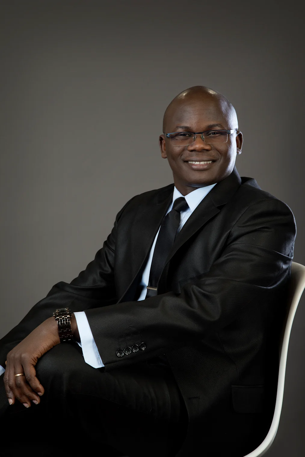 Paul Ekuru, a Black man with a dark skin tone and no hair, sits in a chair. His legs are crossed and he is dressed in a suit. 