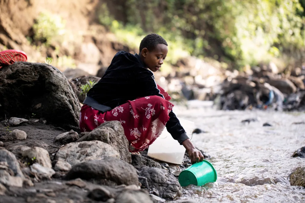 Brenda, a young Tanzanian girl, collects water from a stream using a green bucket. She appears alone and wears a black jacket and red floral skirt.