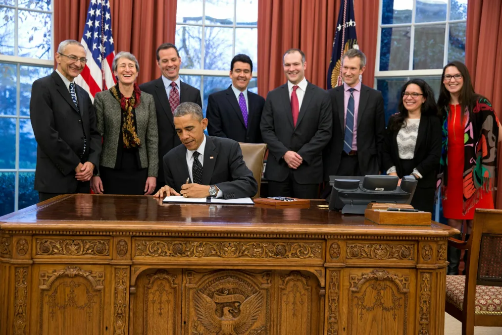 Angela Barranco stands next to seven others as they watch President Obama sign a document in the Oval Office. All people are a range of light to medium skin tones. President Obama sits behind his desk.
