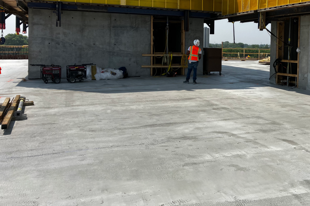 A construction worker in a white hard hat and orange safety vest looks at a structure as he stands on  the concrete floor of Museum Building Level 3