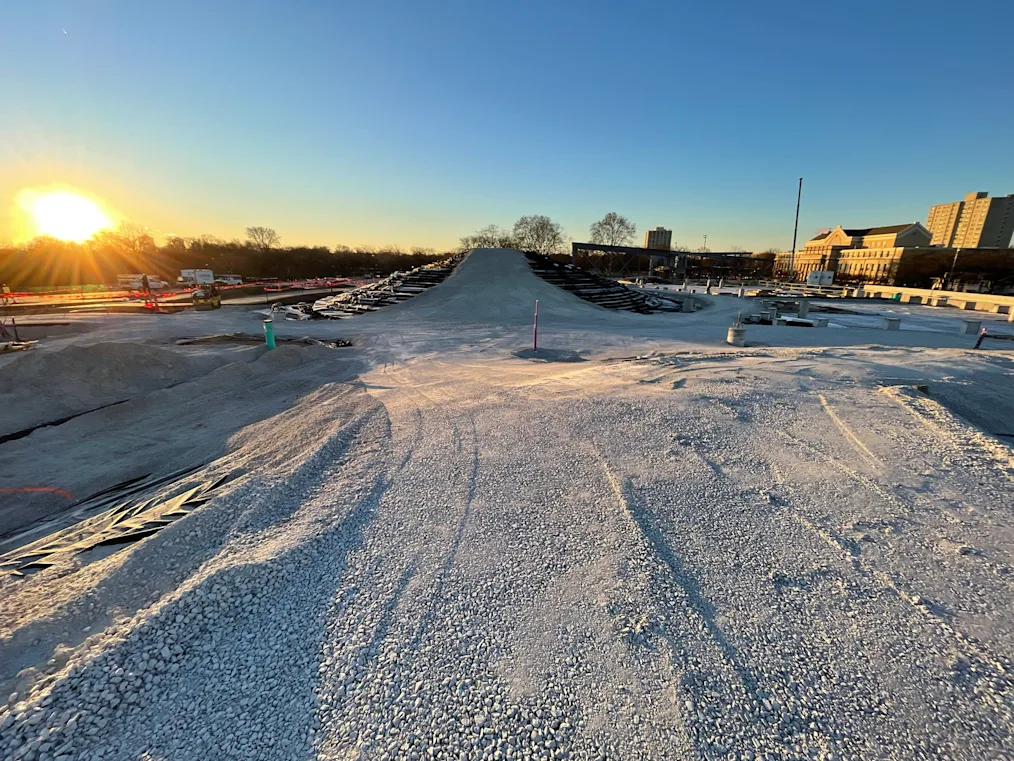 A massive gravel pathway leading up to an incline, meant to one day be a sledding hill. The sun is just rising or setting in the left side of the photo.