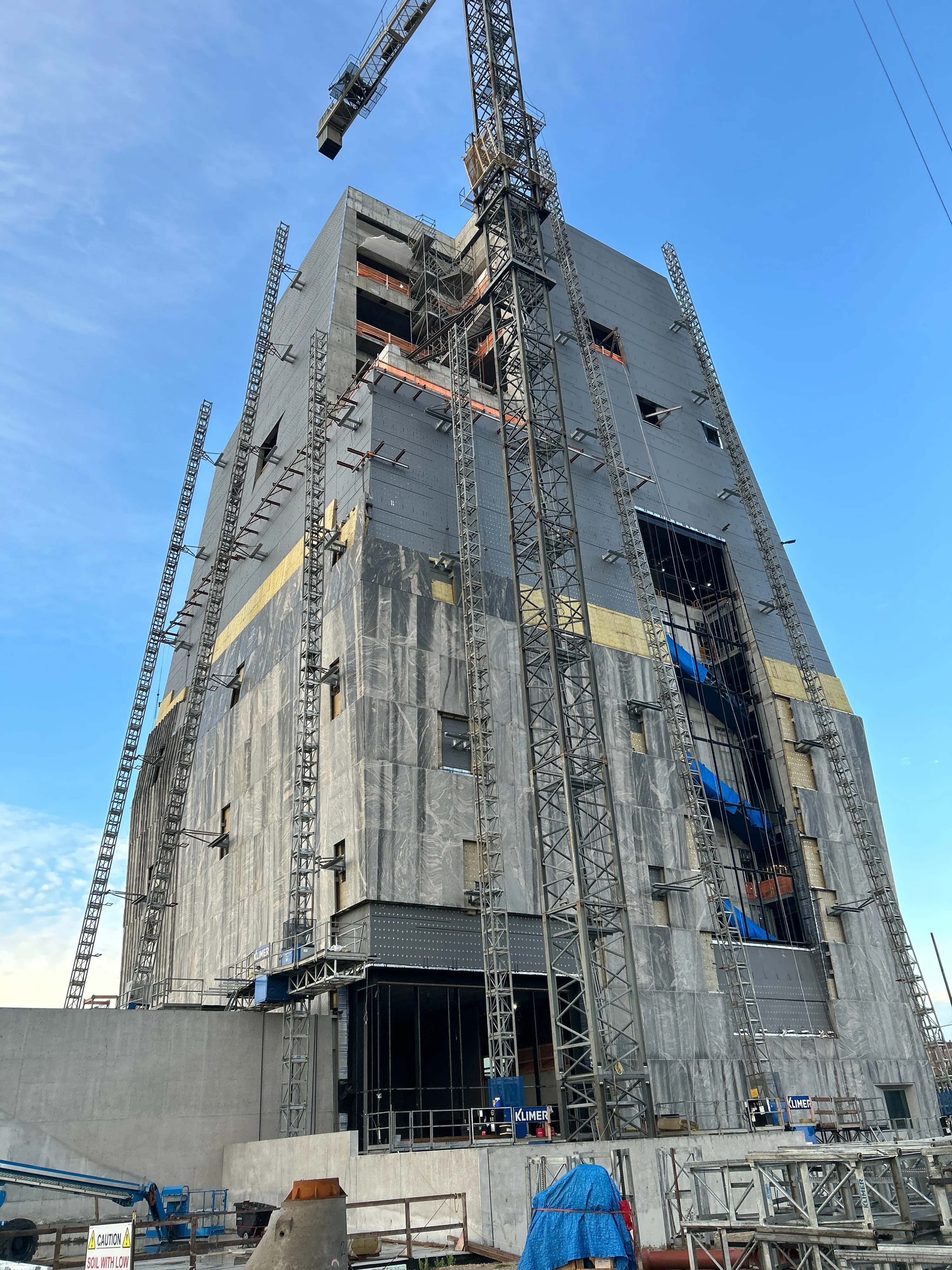 A gray stone installation climbs up the north and east faces of the Museum Building at the Obama Presidential Center. Blue tarp-wrapped escalator bases are seen through an opening that will eventually be clad in an art glass installation. A clear blue sky is in the background.