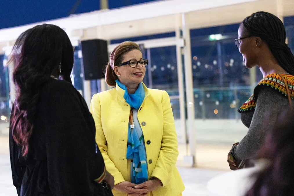 Valerie Jarrett smiles as she stands next to Beverly Ndifoin Niyang, a Black woman with a deep skin tone. She is wearing glasses and braids. Valerie Jarrett is wearing a yellow blazer with a blue scarf.