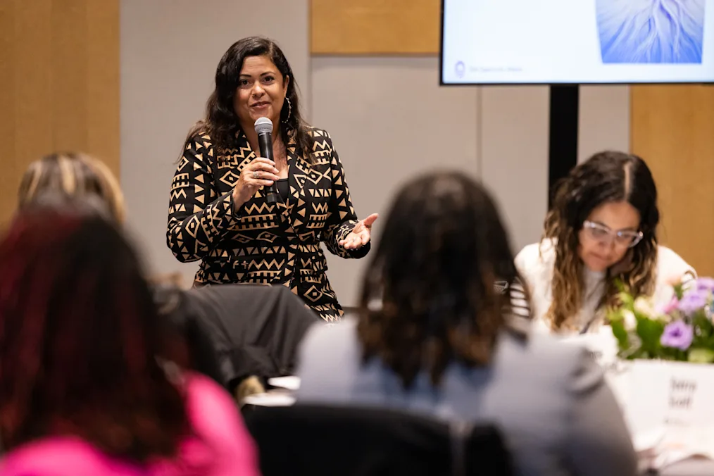 A woman with a medium skin tone speaks into the microphone. She is standing in a room with a group of women with a range of light to dark skin tones. All are dressed professionally. 