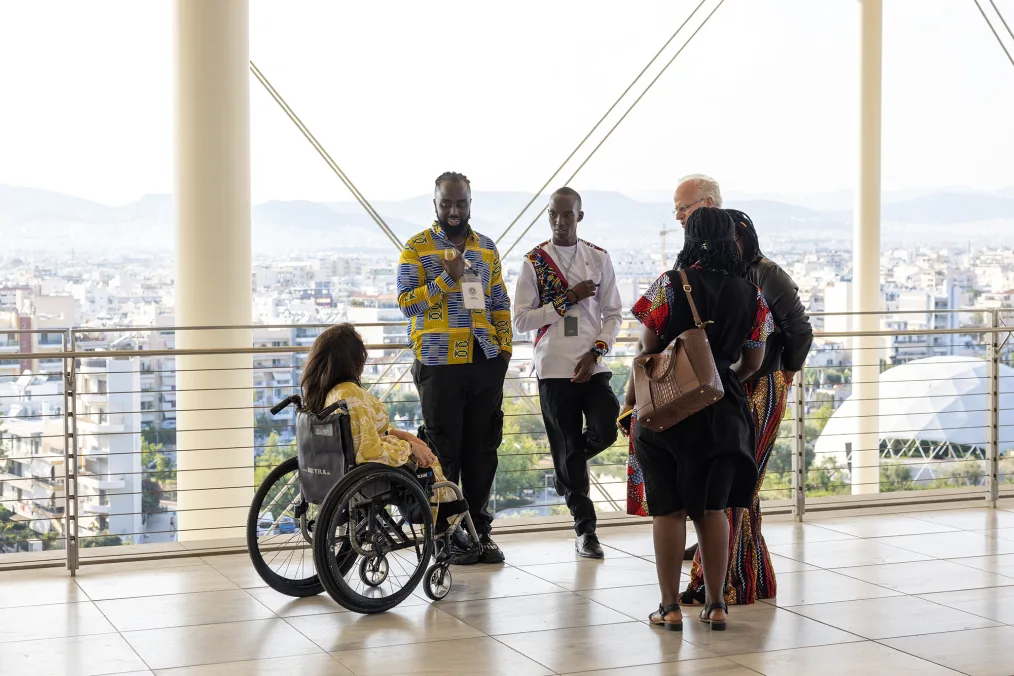A group of people of varying skin tones are talking on the terrace. One woman is using a wheel chair. In the background is the city of Athens.