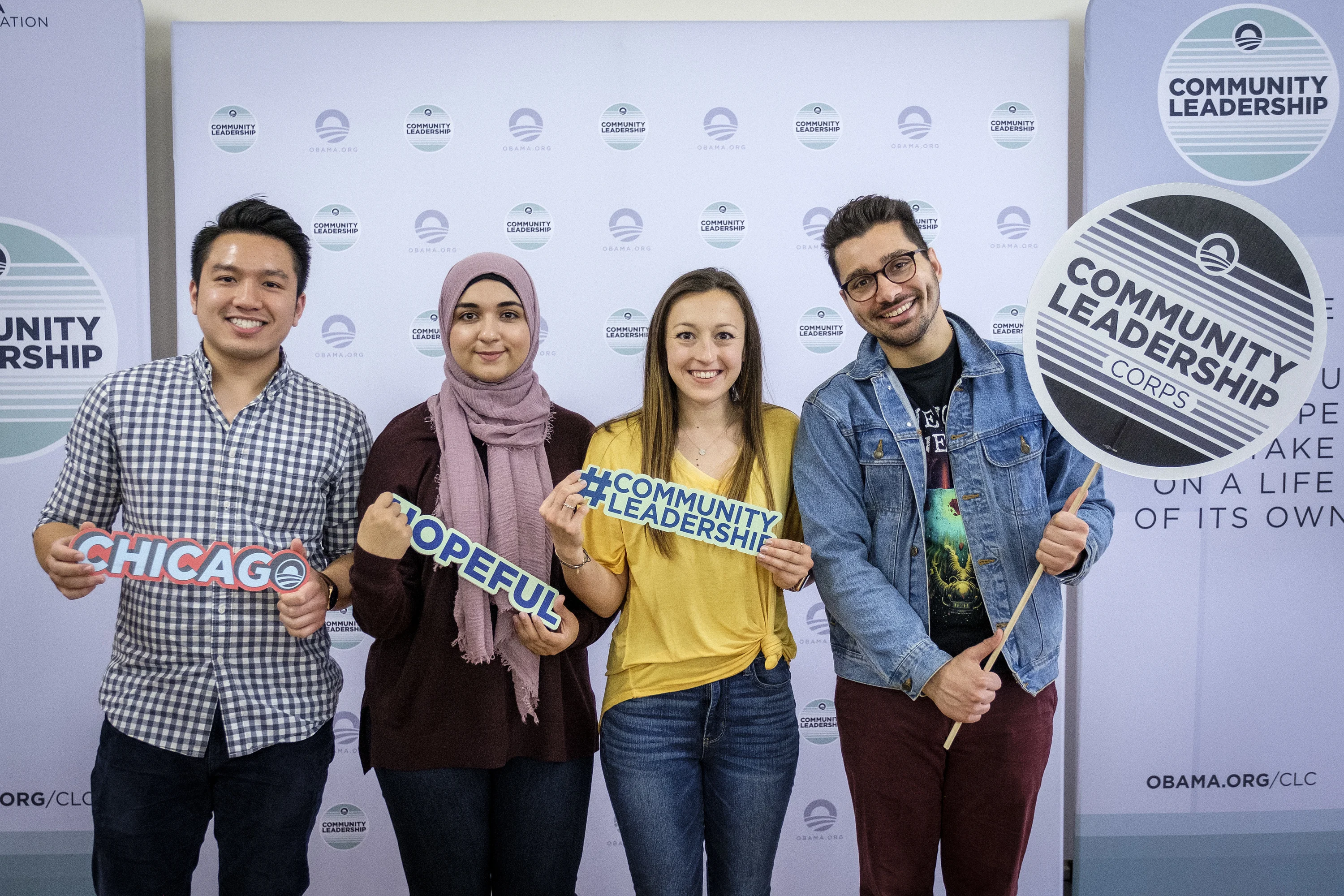 Four individuals with light skin tones stand in front of a blue Obama Foundation backdrop holding signs thats say "community leadership" and "hopeful". 