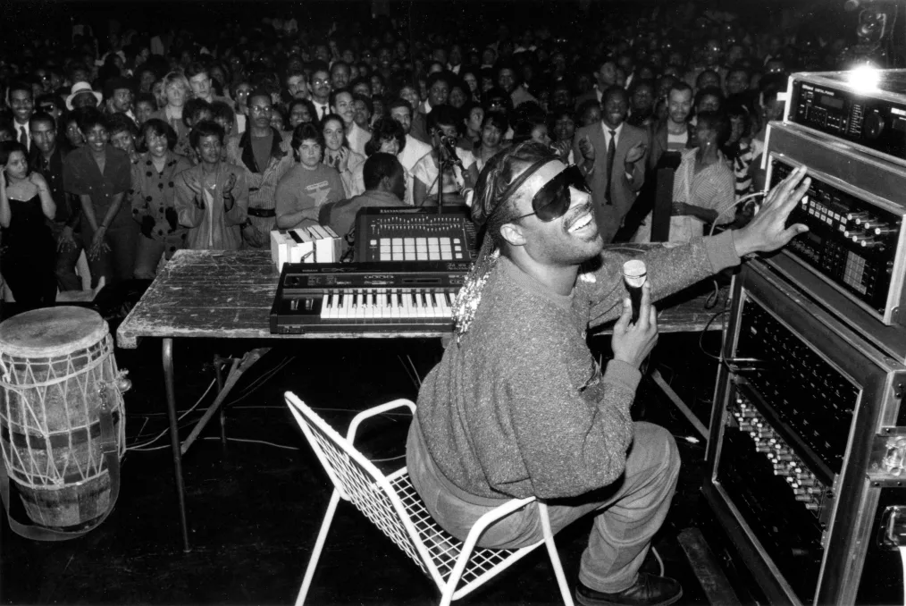 A black-and-white image shows a man with a deep skin tone with braids and special glasses sitting in
a chair with a microphone playing the keyboard and adjusting the speaker volume.