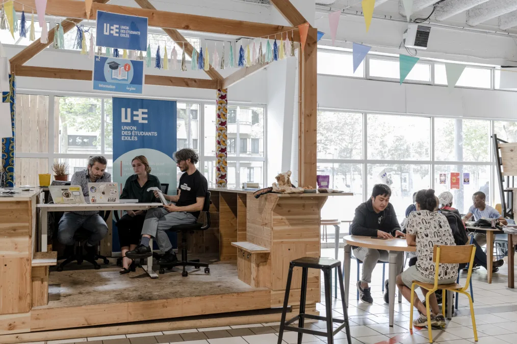 There is a well-lit space with white walls and windows with a brown wooden section with steps to enter. There are three people with a skin tones sit at a small table on the brown platform and other individuals of different skin tones sitting outside of it. 