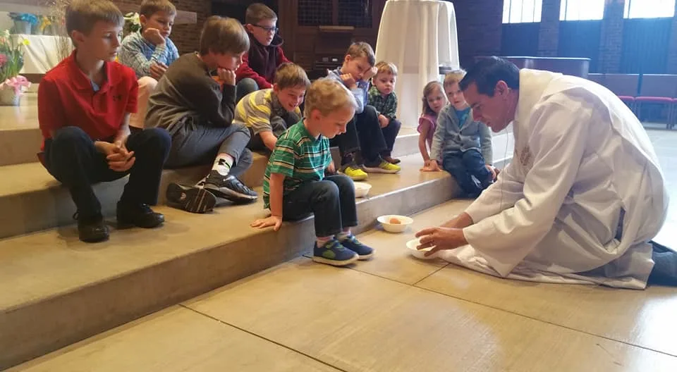 Jerad Morey, a man with a light skin tone and salt and pepper hair, kneels to grab an egg in a plastic bowl. He is surrounded by a group of young children with light skin tones and multiple ages. 