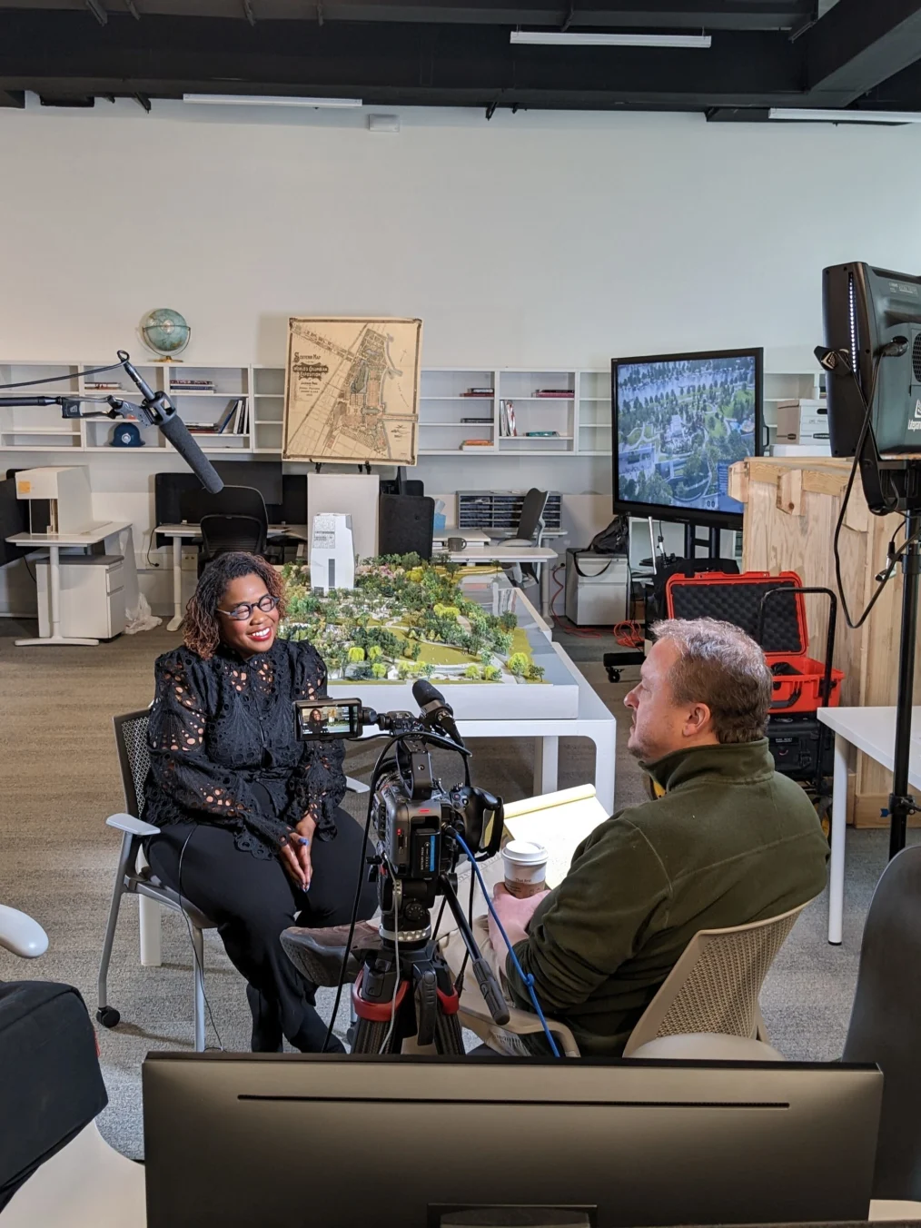 In this photo, Crystal Moten sits in a chair across from Tom Steffus, a white man with a light skin tone and thin brown hair, for an interview about the Obama Presidential Center. An office space and 3D model of the Obama Presidential Center is in the background. 