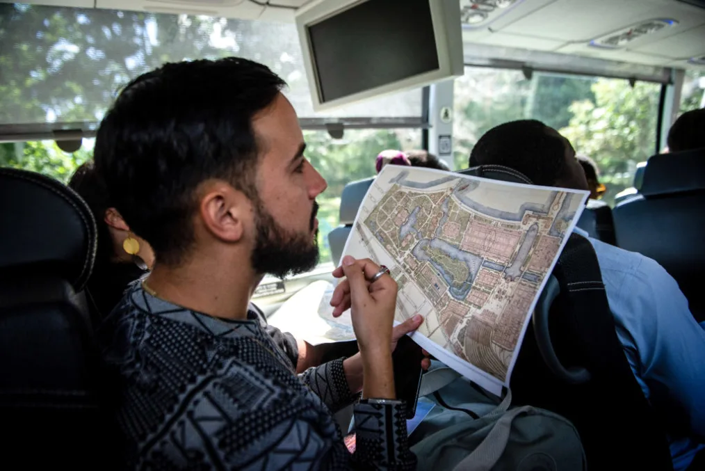 A group of people riding on a tour bus. One person is in focus in the center of the photo. It is a man wiht short full, slick black hair, a full beard aswell. He is wearing a feint blue swearter with balck patterns on it. In his lap is a map of the OPC park, a phone, and a travel bag.