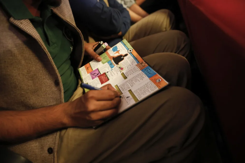A man wearing a dark beige flannel suit jacket, green polo shirt, and beige pants wries in a book that has the colors blue, red, orange, and pink inside. the pictures on the book depict wellness.