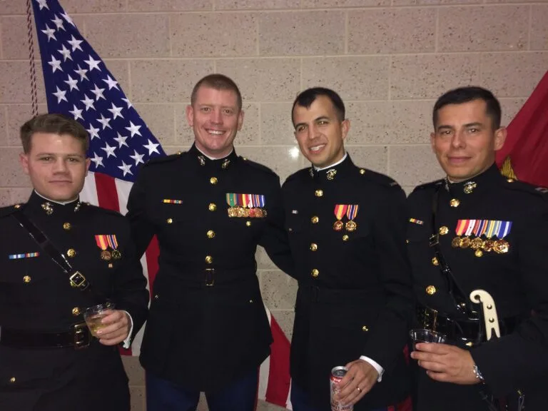 Four men in Marine blue dress uniform smile and pose for a photo in front of an American and Marine flag.