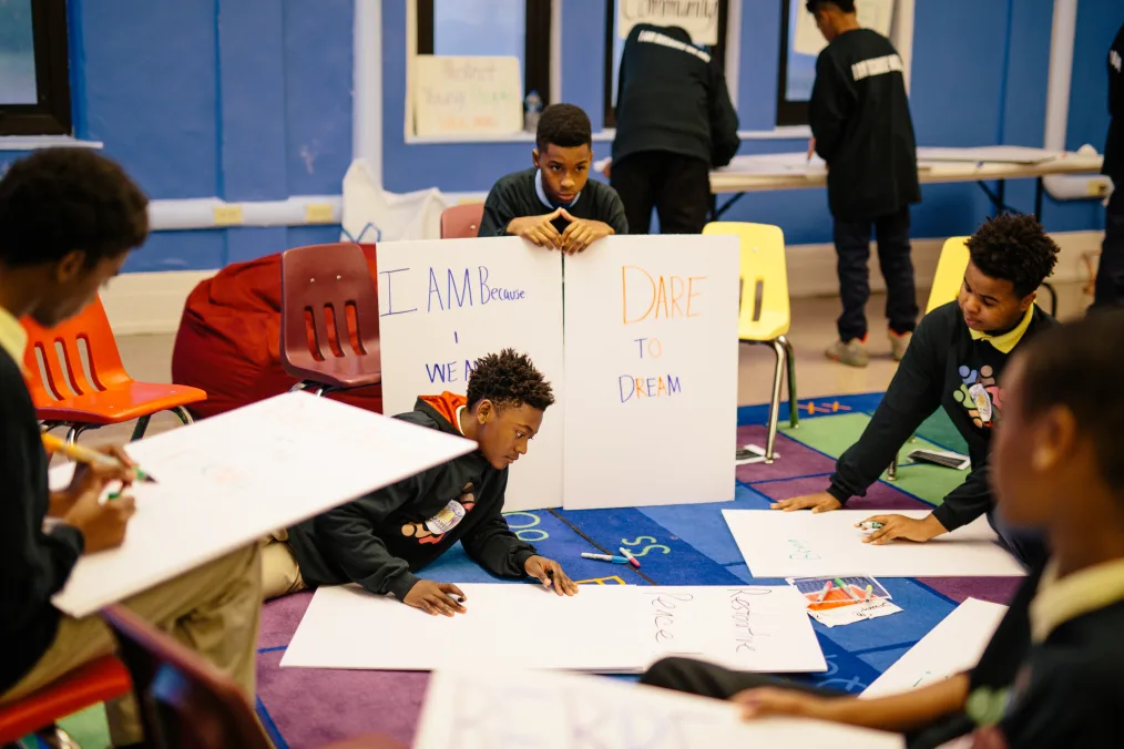 Seven young men in uniforms with a range of light to deep skin tones create posters. The boy in the center has a medium skin tone and is holding two signs that read, “I am because we are'' and “Dare to dream.”