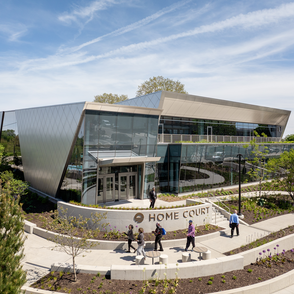 A wide high-angle view of a modern two-story building labeled "HOME COURT." It features large glass windows and a geometric silver metal roof. A low curved concrete wall in front displays the text. Several people walk on light-colored concrete paths surrounding the building, which is bordered by mulch beds and green plants under a clear blue sky.