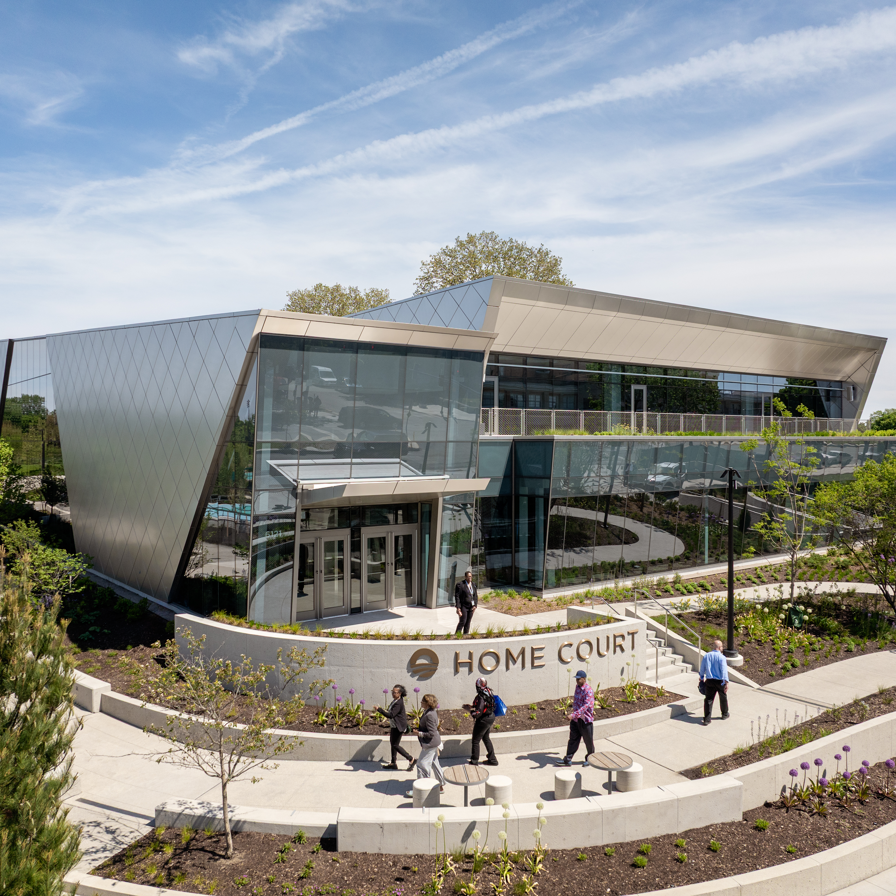 A wide high-angle view of a modern two-story building labeled "HOME COURT." It features large glass windows and a geometric silver metal roof. A low curved concrete wall in front displays the text. Several people walk on light-colored concrete paths surrounding the building, which is bordered by mulch beds and green plants under a clear blue sky.