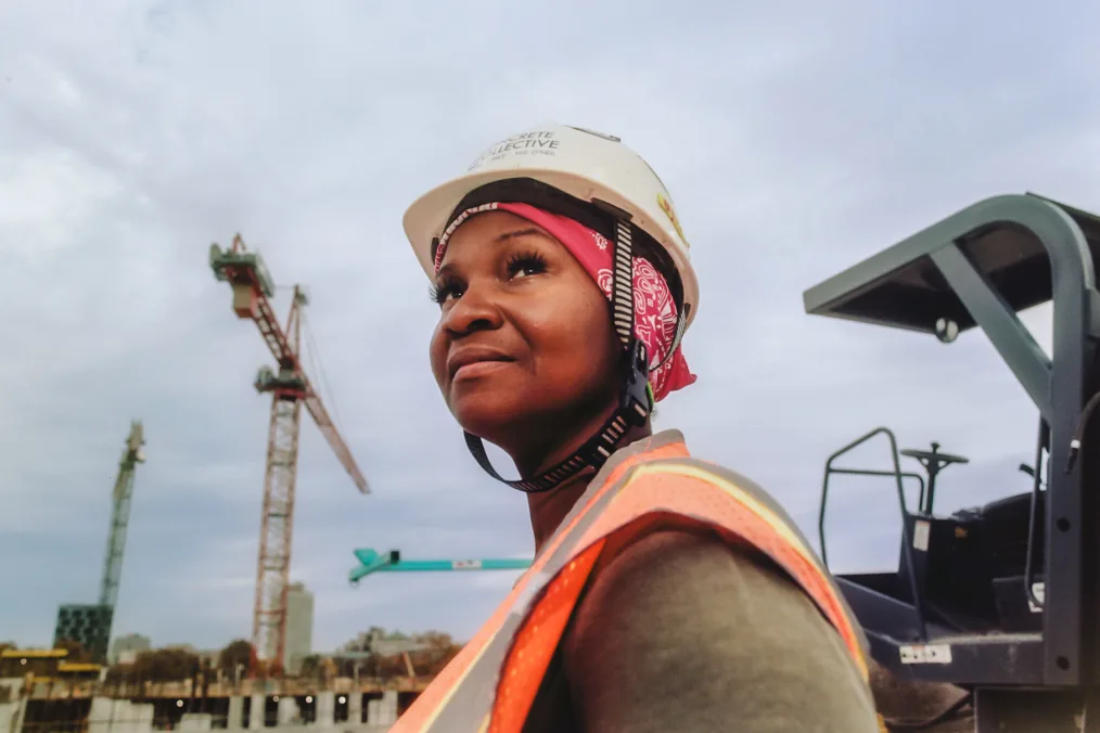 A woman with a medium-deep skin tone wearing an orange, gray, and yellow construction jacket and helmet looks up toward the sky while on a construction site. There are cranes in the background. 