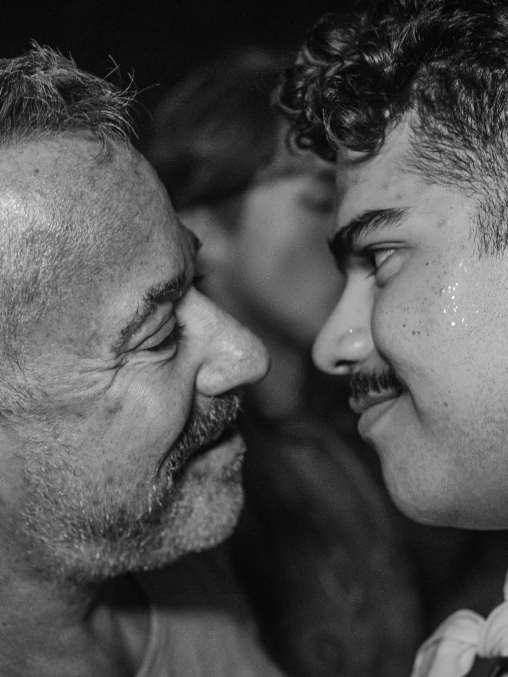 The image is a photo of two people close together in the middle of an embrace staring at each other. The photo is a closeup in black and white. The first person has short close cut hair and a goatee. The second person has short dark curly hair, a dark mustache, and a bandana tied around their neck.